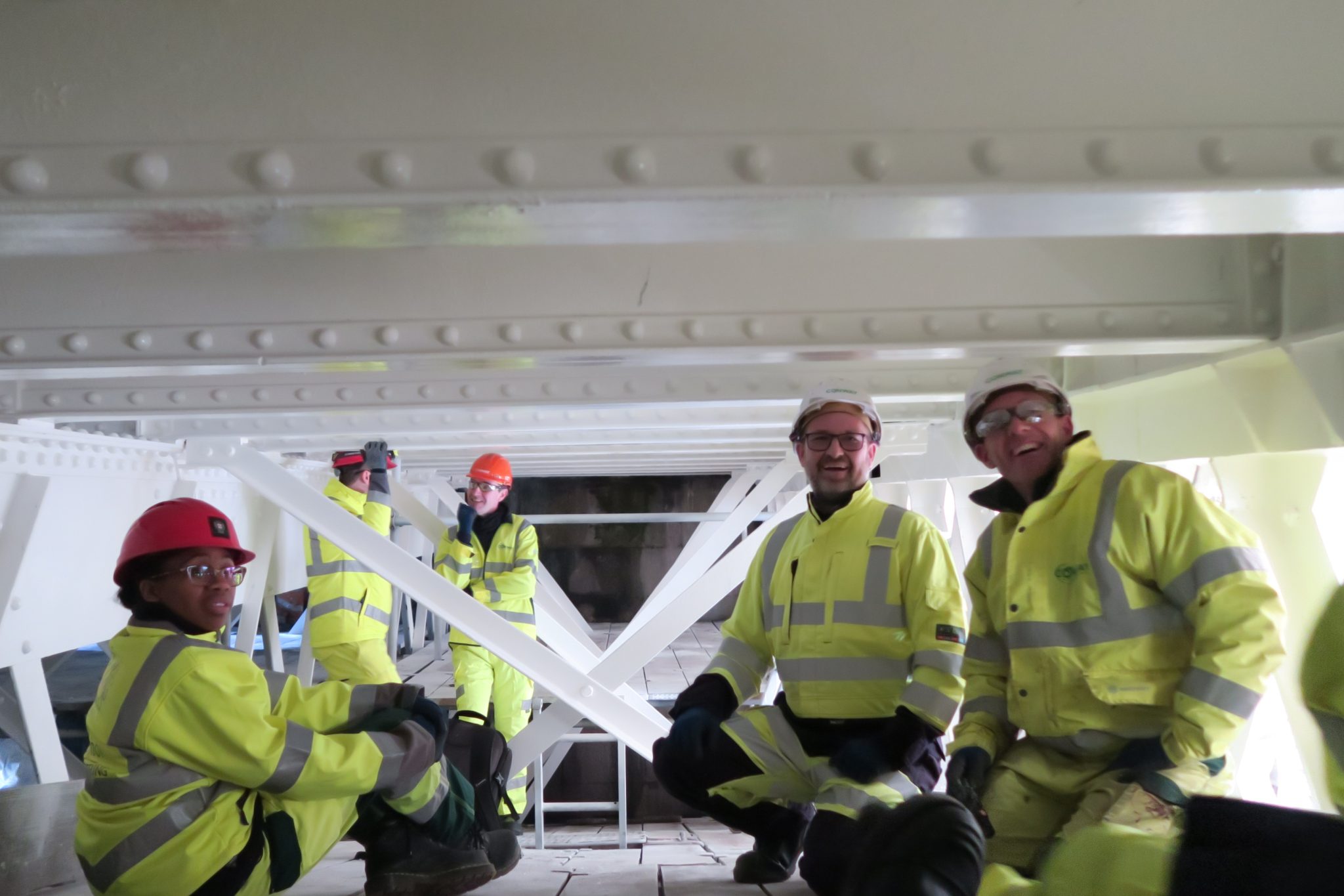 Five people wearing bright yellow personal protection equipment and helmets explore a cramped environment of white girders and metalwork.