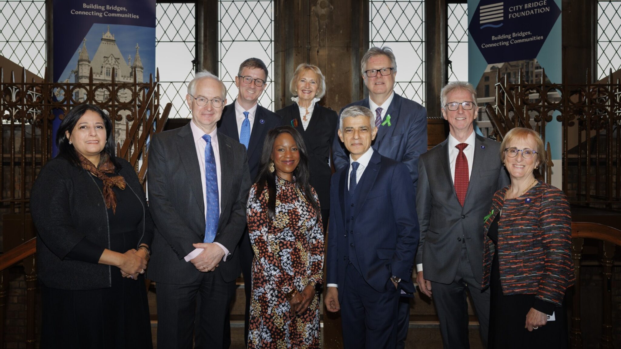 A photo taken at a City Bridge Foundation's annual board dinner inside Tower Bridge. It shows a group of nine people, including City Bridge Foundation Managing Director, Board Members and The Mayor of London and Deputy Mayor for Communities and Social Justice. Two City Bridge Foundation banners visible in the background displaying the text “City Bridge Foundation – Building Bridges, Connecting Communities.”