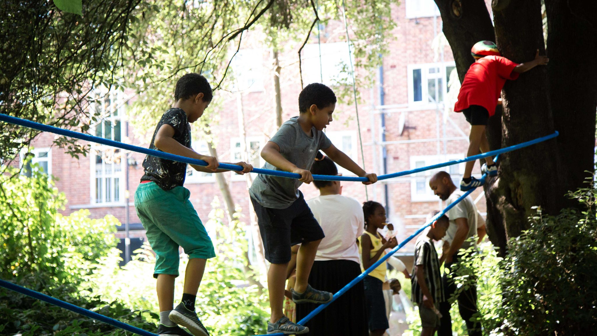 Children playing on a basic rope walk strung between two trees, under a natural canopy of thick leaves. Housing can be seen in the background.