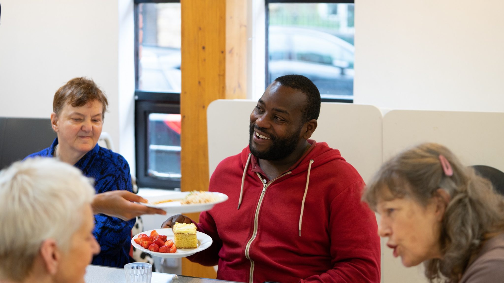 A smiling man accepts a place of cake and strawberries in a community cafe. He is sitting at a table with other people who are chatting to each other.