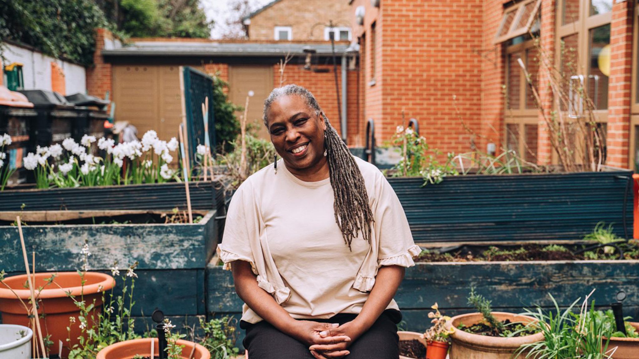 A smiling Black woman sits amid vegetable beds, planters and pots, in a community garden.