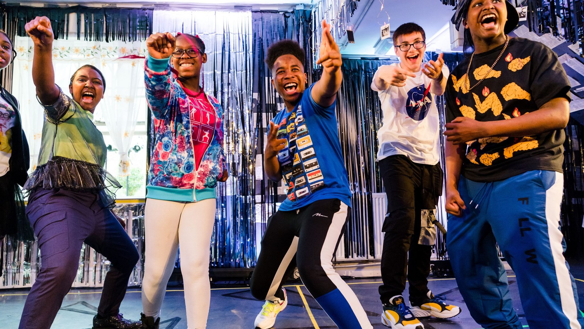Five young people joyfully celebrating on a colourful stage at Kiln Theatre Company. Image credit: Alex Brenner