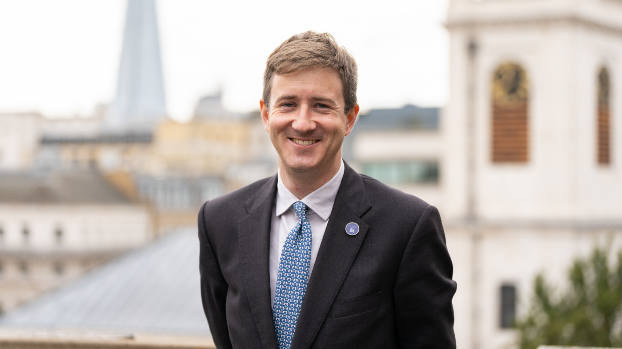 A smartly dressed man in a suit jacket with a blue tie is smiling. Behind him can be seen London rooftops and the Shard.