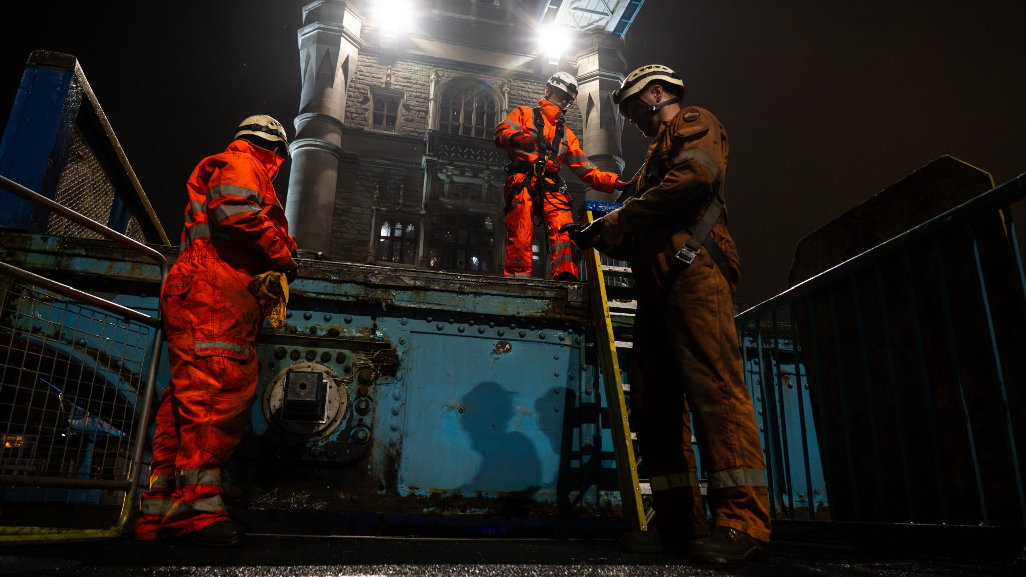 Three engineers in full protective gear and hard hats work on Tower Bridge at night.
