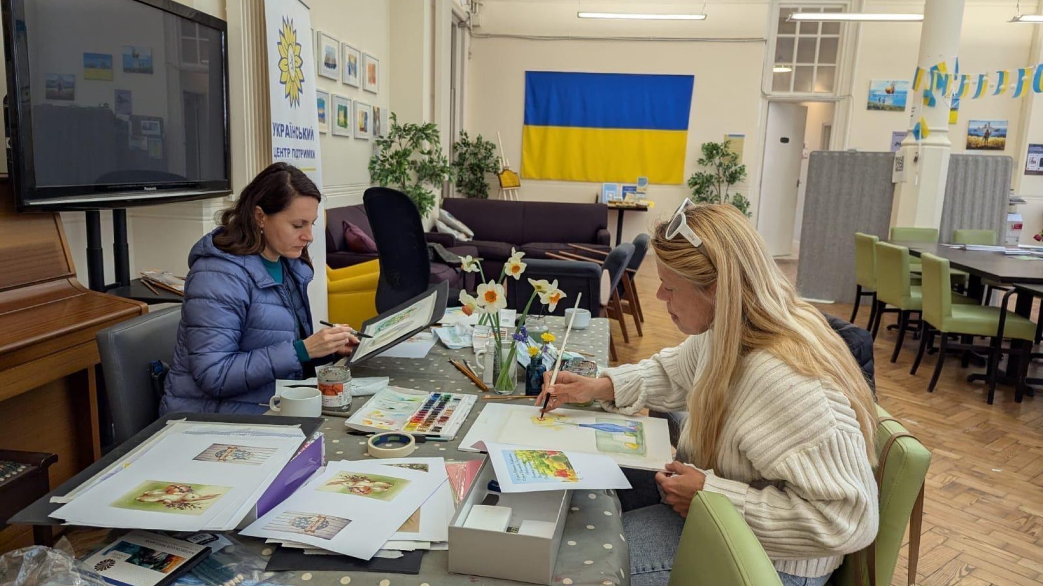 Two women sitting at a table taking part in art activities. The Ukrainian flag is displayed on the wall in the background.