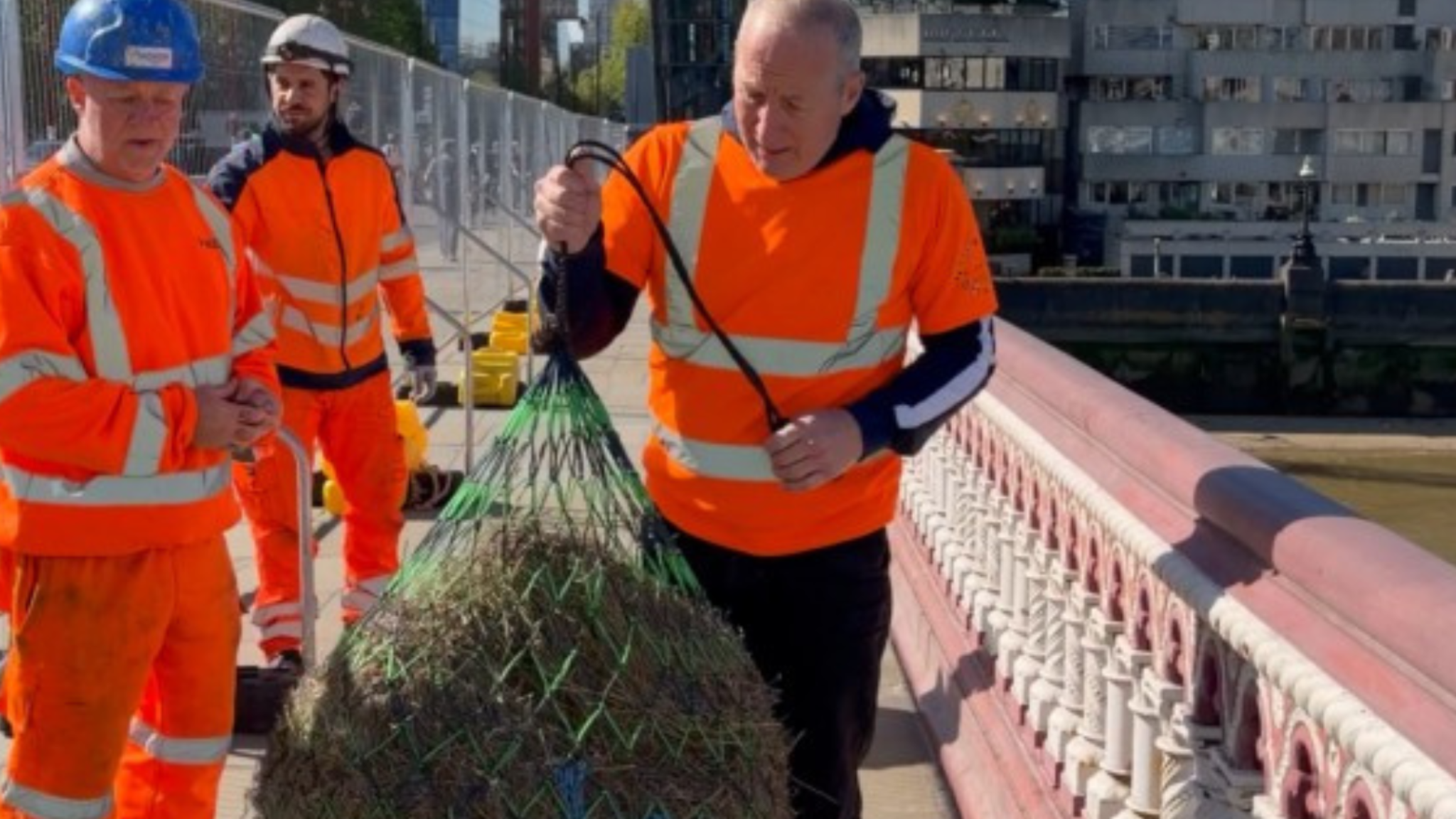 A man dressed in bright personal protective clothing holds a large bundle of straw next to the edge of Blackfriars Bridge.