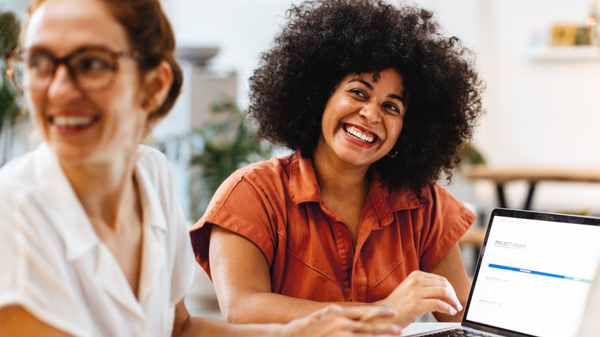 Two women working at an office laptop and smiling cheerfully at someone else.